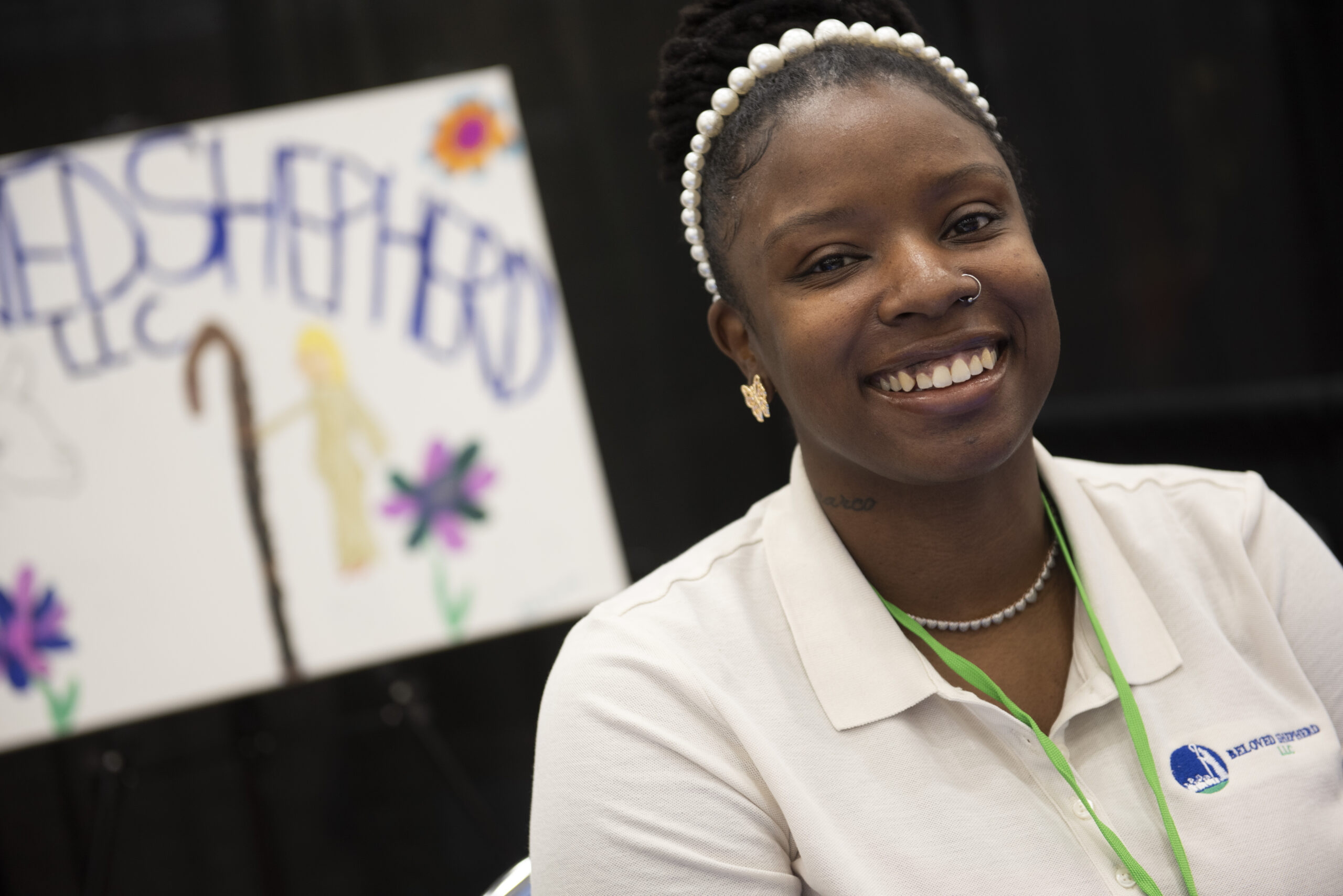 A black woman smiles at the camera. She wears a pearl headband and a green lanyard over her white collared shirt.